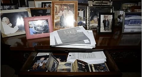 A collection of framed family photos and paperwork clutter a wooden desk. An open drawer reveals more photographs. The scene feels nostalgic and personal.