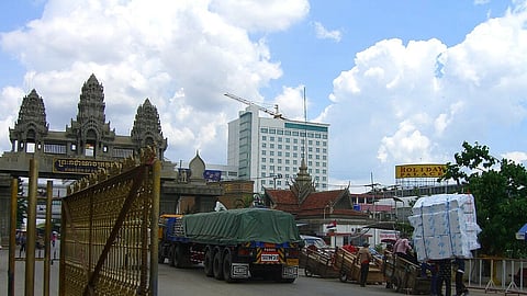 A bustling border crossing features ornate stone gates with a large truck passing through. People with carts and large packages move nearby under a sky with fluffy clouds.