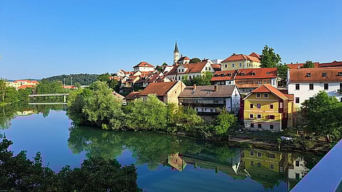 Charming riverside village with colorful houses, lush greenery, and a tall church spire under a clear blue sky reflected in calm water. Tranquil scene.
