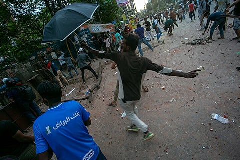 The image depicts a riot broken out in Bangladesh, with a man wearing a mask and holding an umbrella, is throwing a bottle. People all around are involved in riots.