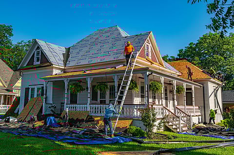 Workers repair the roof of a Victorian-style house. A man in an orange shirt stands on a ladder, while others work on the roof. Sunny day, lush greenery.