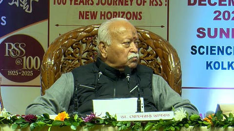 Mohan Bhagwat sits on an ornate chair at an event, featuring floral decorations and a microphone. Background shows signs: "100 Years Journey of RSS," "December 2025," "Kolkata." His expression is thoughtful.