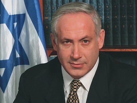 Portrait of Israeli PM Benjamin Netanyahu. he is wearing a black suit and behind him is the flag of Israel and shelf filled with books.
