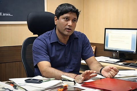 Sanjiv Chaturvedi in a blue shirt sits at an office desk, gesturing as he speaks. Papers and a phone are on the desk, with a computer screen in the background.