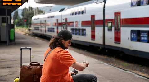 A person in an orange shirt and cap checks their watch while seated on a train platform. A suitcase is next to them, and a train is in the background.