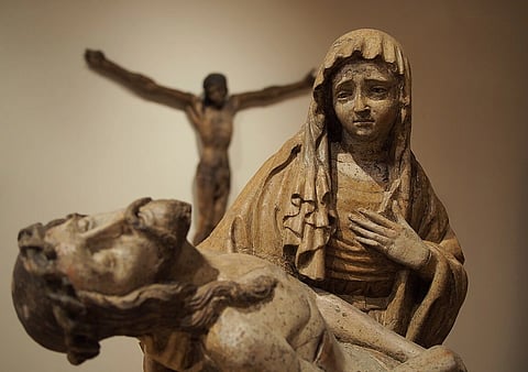 A sorrowful sculpture depicts a grieving woman cradling a lifeless figure in front of a blurred crucifix figure, conveying deep sadness and compassion. Sculpture of Crucifixion of Jesus observing Virgin Mary cradling the dead body of Jesus (known as Pieta). National Gallery of Slovenia. 14th and 15th century.