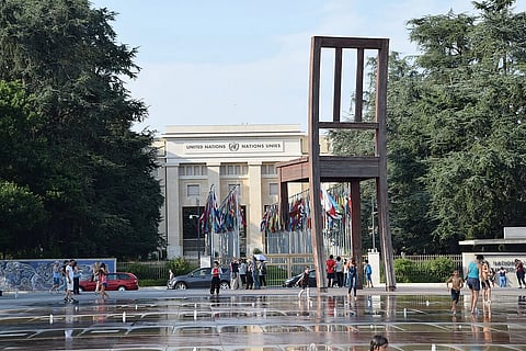 The image depicts United Nations Building, Geneva. Flags of multiple nations are in front of the building, and people are walking outside.