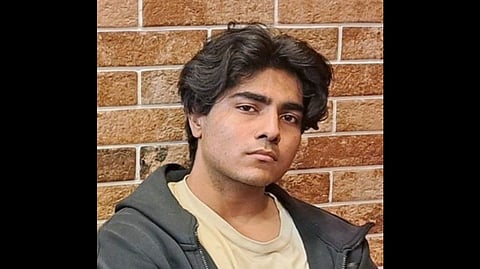 Indian Doctoral student Shivank Avasthi with wavy dark hair and neutral expression, wearing a gray hoodie over a beige shirt; stands against a textured brick wall.