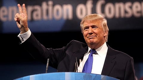 In the image Donald Trump is shown wearing a coat with blue tie he is showing a victory sign and smiling at the people