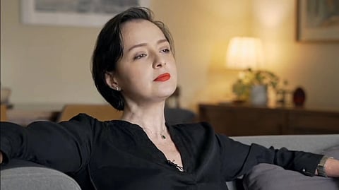 A woman with short dark hair leans back on a sofa, wearing a black shirt and red lipstick. The room is softly lit, creating a calm, reflective atmosphere.