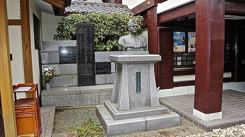 A bronze bust of Subhas Chandra Bose on a stone pedestal is placed in a courtyard. Behind it are polished stone plaques with inscriptions, and flowers are arranged nearby. The setting is serene and respectful.