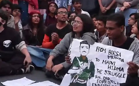 The image depicts a group of protesters protesting against the Air pollution crisis at India Gate, New Delhi. They are holding a poster that mentions: "From Birsa Munda to Madvi Hidma, The Struggle for our forests and environment", the rest is out of the frame.