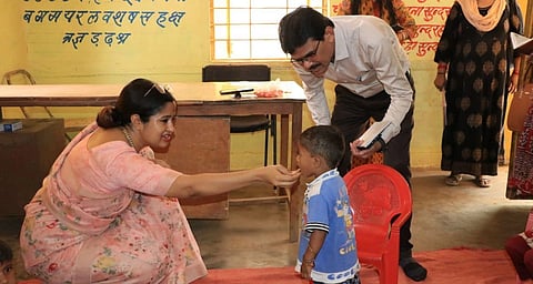 A woman in a pink saari crouches down to a feed a small child with her hand while a man in a white shirt stands behind the child.