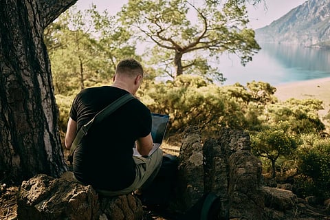 A man sits with a laptopn on a rock in the middle of a forest. There are mountains and a lake in the background.