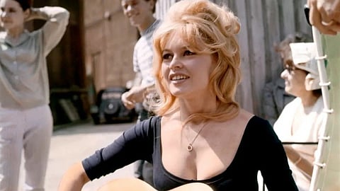 Legendary French Actress Brigitte Bardot with blonde hair plays an acoustic guitar, smiling warmly. She is outdoors with three people in the background, exuding a joyful and relaxed atmosphere.