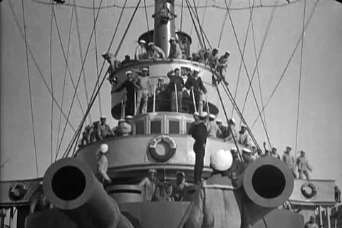 Black and white photo of sailors on a ship's deck, crowded around the structure, ropes extending upward, creating a bustling, dynamic scene.