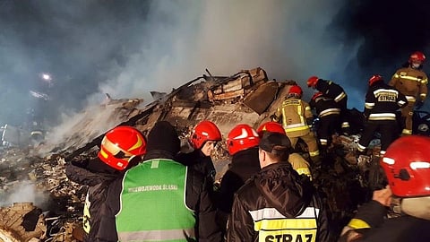 Firefighters in red helmets and uniforms, labeled "STRAŻ," work amidst smoke and rubble at night, conveying urgency and determination in a disaster scene.