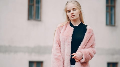Young woman with long blonde hair wearing a stylish pink fur coat and black top stands confidently outside with a blurred building in the background.