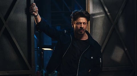Bollywood actor SRK in dark casual attire leans confidently against a lit doorway in a dim, industrial room setting