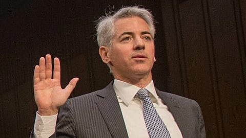 In the image Bill Ackman is shown raising his one hand he is wearing formal suit behind looks like professional setting
