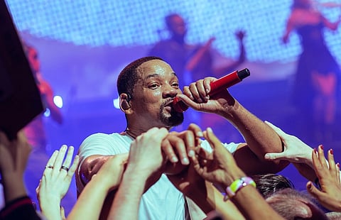 Image of actor rapper Will Smith doing a concert with a crowd holding his hand. he is holding a mic in his another hand. the image is bluish tone.