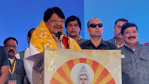 Kailash Vijayvargiya is speaking at event, surrounded by people behind him. He is speaking at a podium, wearing an orange gamcha and a white kurta.