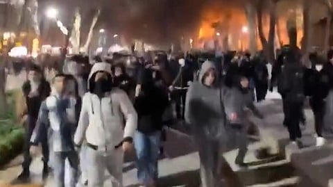 A large crowd of people walks through a dimly lit street at night with trees on either side as they protest in Bangladesh. The scene is tense with an orange glow in the distance.