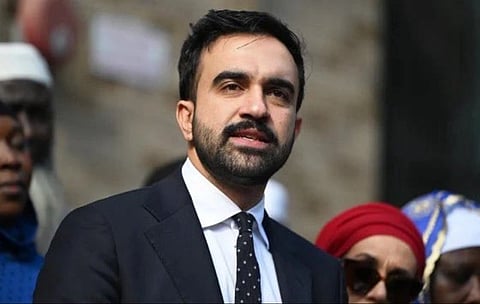 In the image Zohran Mamdani is shown adressing people he is wearing black coat looking at crowd