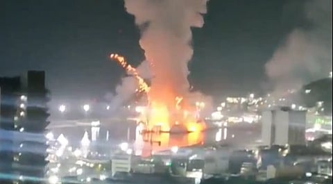 Nighttime scene of an explosion on water near a city, with towering flames and smoke illuminating the surroundings, creating a dramatic and urgent atmosphere.