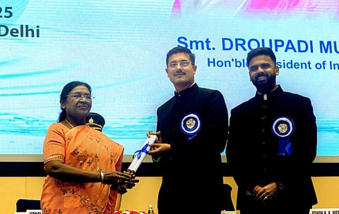 Image of president Droupadi Murmu giving award to 2 men. She is handing over a certificate tied in blue ribbon to a man in a black suit with a blue emblem. Another man in a similar suit stands smiling. Background displays text, indicating an official event.