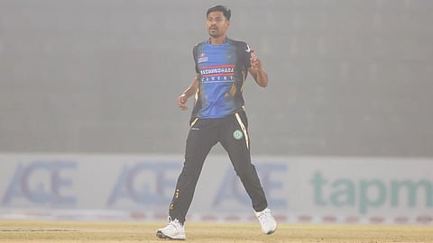 Bangladesh cricketer Mustafizur Rahman wearing a blue and black jersey stands on a foggy field, mid-action, appearing focused. Advertising boards are blurred in the background