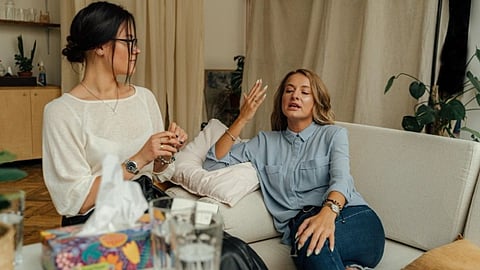 Two women are sitting on a couch having a conversation. One gestures passionately. The atmosphere is relaxed, with plants and a colorful tissue box.