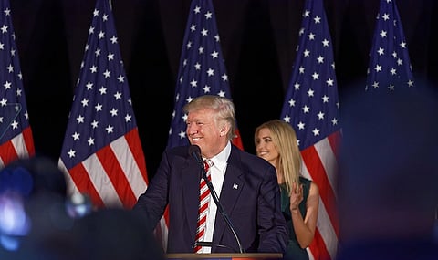 Image of Donald Trump at Aston. 5 American flags are visible in the background. trump is looking towards his right while giving a speech in front of a podium.