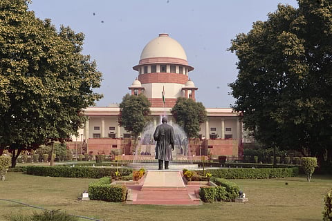 The image shows the Supreme Court of India, with India's flag in front, a lawyer's statue and greenery all around.