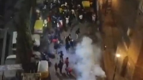 Aerial view of a chaotic street scene at night, with a crowd scattering amid thick smoke and overturned barriers at the Turkman Gate area of Central Delhi, conveying tension and urgency.