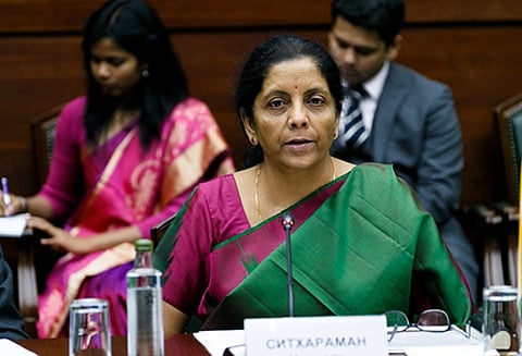 Image of FM Nirmala Sitharaman in a green and pink sari speaks at a conference table, looking attentive. Two people take notes behind her,