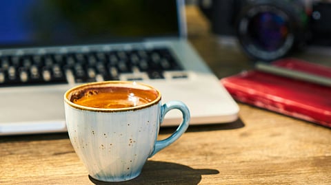 A steaming cup of espresso in a light blue mug sits on a wooden table. In the background are a partially visible laptop and a vintage camera.