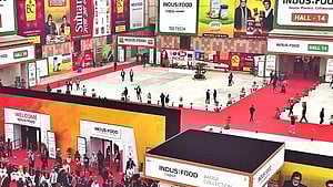 A bustling trade show with bright red carpets, numerous stalls, and large advertising banners. Crowds of people walk and gather around exhibits, creating a lively atmosphere.