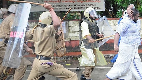 Police officers in riot gear running after a man in a lungi and beating him with lathis.