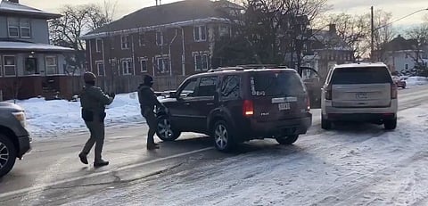Two officers wearing tactical gear approach a stationary SUV on a snowy residential street. The scene appears tense against a backdrop of winter houses.