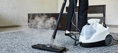 A person using a steam cleaner on a gray carpet in a modern bedroom. The steam is visible, and the atmosphere is clean and organized.