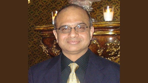 A man with glasses and a pleasant expression wears a suit and striped tie, standing in front of an ornate wooden mantel with lit candles, creating a warm ambiance.