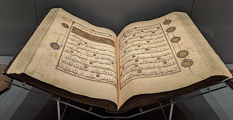 Ancient open manuscript with Arabic calligraphy on yellowed pages, adorned with intricate gold and blue floral motifs, displayed on a stand.