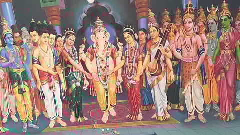 A vibrant painting depicts Hindu deity Ganesha in the center, surrounded by other colorful deities in traditional attire, conveying a festive, sacred atmosphere.