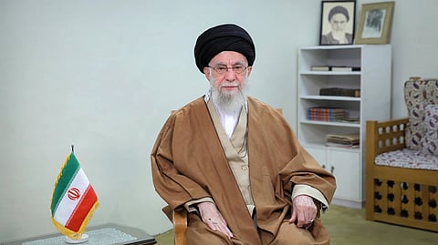 Iran's Supreme leader Supreme Leader Khamenei in traditional brown attire sits in a modest room. An Iranian flag is on a small table beside him. Books and photos are on a shelf behind.