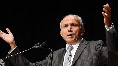 Prem Watsa in a suit raises his arms while speaking at a podium with microphones. His expression appears engaged, set against a dark background.