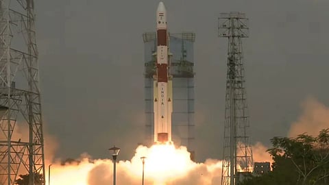 A rocket labeled "India" launches amid billowing smoke and bright flames against a gray sky. Tall structures frame the scene, conveying power and excitement.