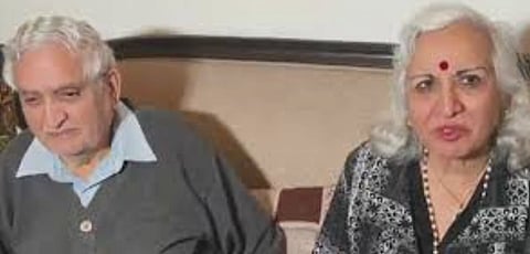 Image of two elderly couple. The man wears a gray sweater over a blue shirt, while the woman has white hair, a red bindi, and a patterned blouse. Warm ambiance.