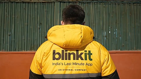 A person in a yellow jacket with "Blinkit: India's Last Minute App" stands against a teal corrugated background. The scene conveys efficiency and modernity.