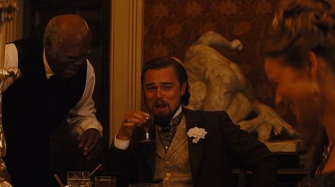 In the image a movie scene from Django Unchained is shown in which leonardo is holding a wine glass and there are two other people, the background is vintage